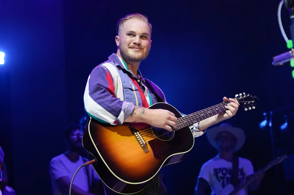 Zach Bryan performing on his American Heartbreak Tour, holding an acoustic guitar and wearing a multi-colored striped shirt.