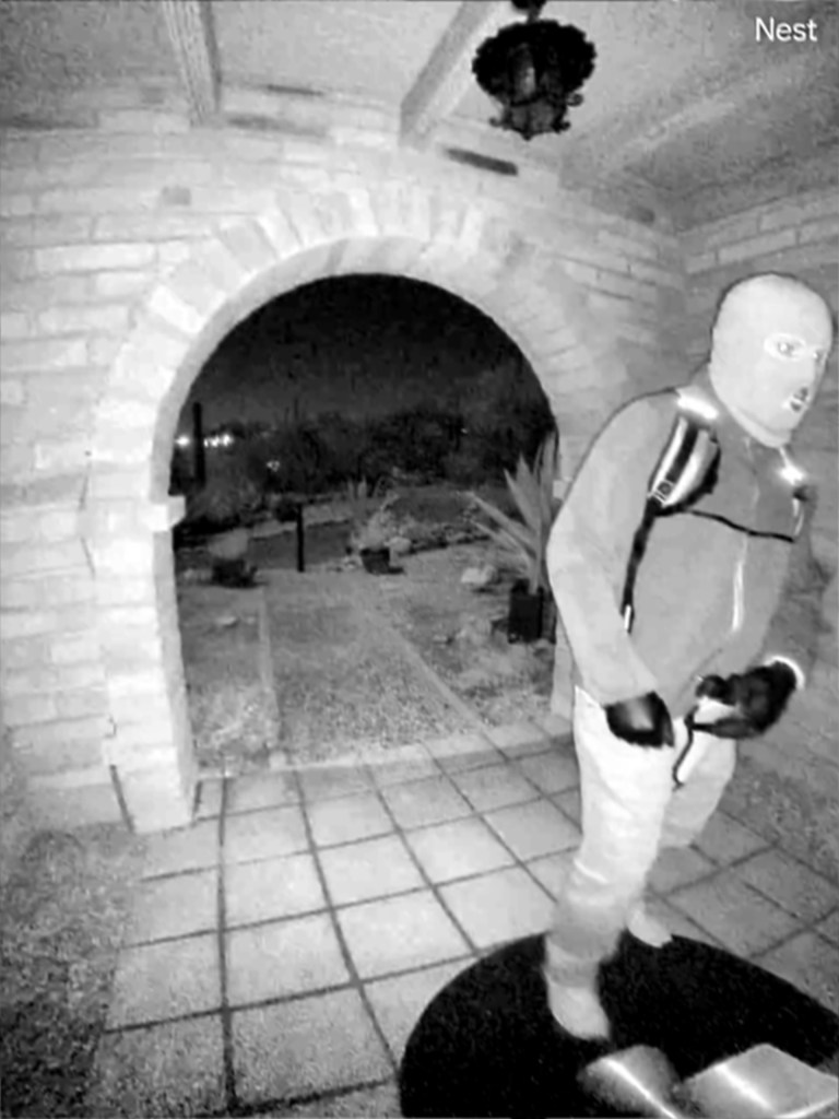 Surveillance image of a person in a balaclava, jacket, and backpack at a doorway.