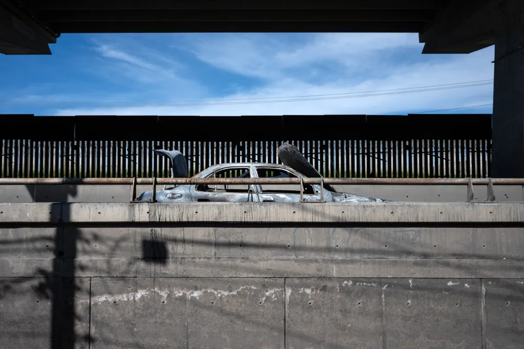 A burned car sits on a concrete barrier under a bridge, with a metal fence and blue sky in the background.