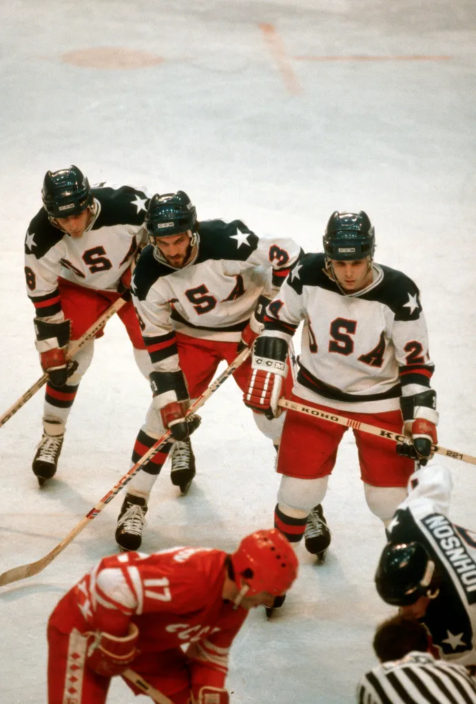 Three US hockey players in white, navy, and red jerseys face a Soviet player in a red jersey and a referee in a striped shirt during a game at the 1980 Winter Olympics.