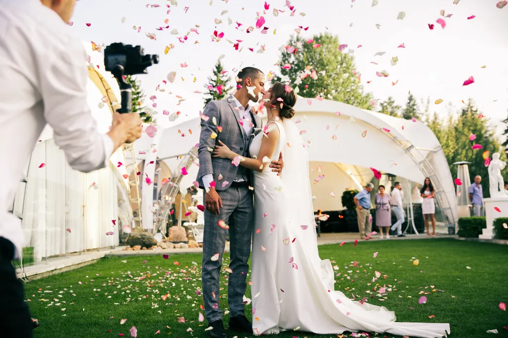 A photographer captures a newlywed couple kissing as rose petals fall around them on their wedding day.