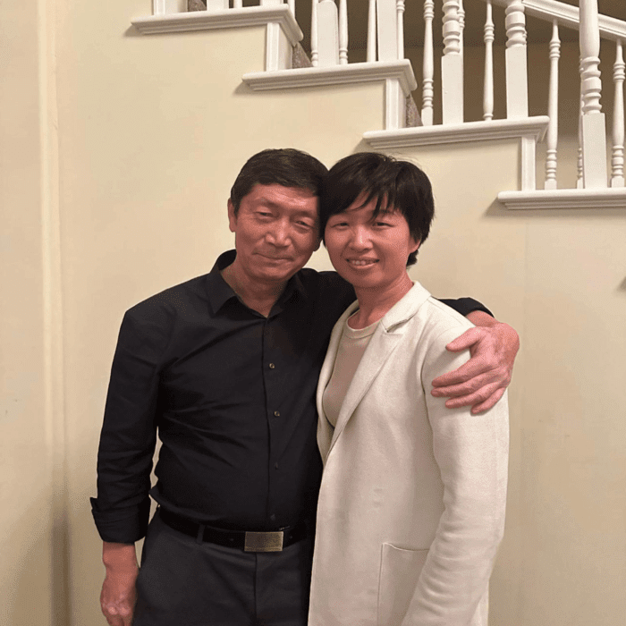 A man in a black shirt and a woman in a white blazer smiling with an arm around each other's shoulders.