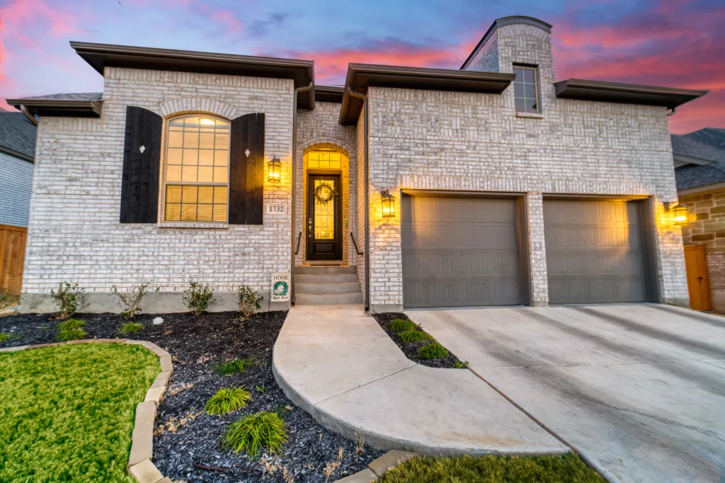 A white brick home with yellow windows and a large driveway, at twilight under a pink and purple sky.