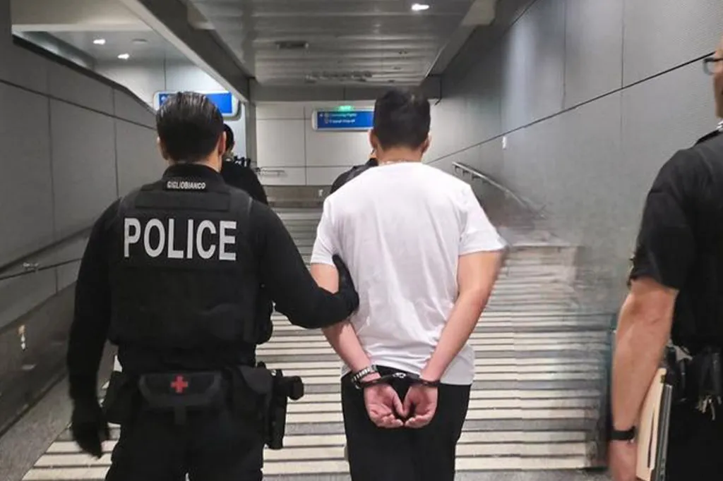 Police escort a man in handcuffs down a hallway.