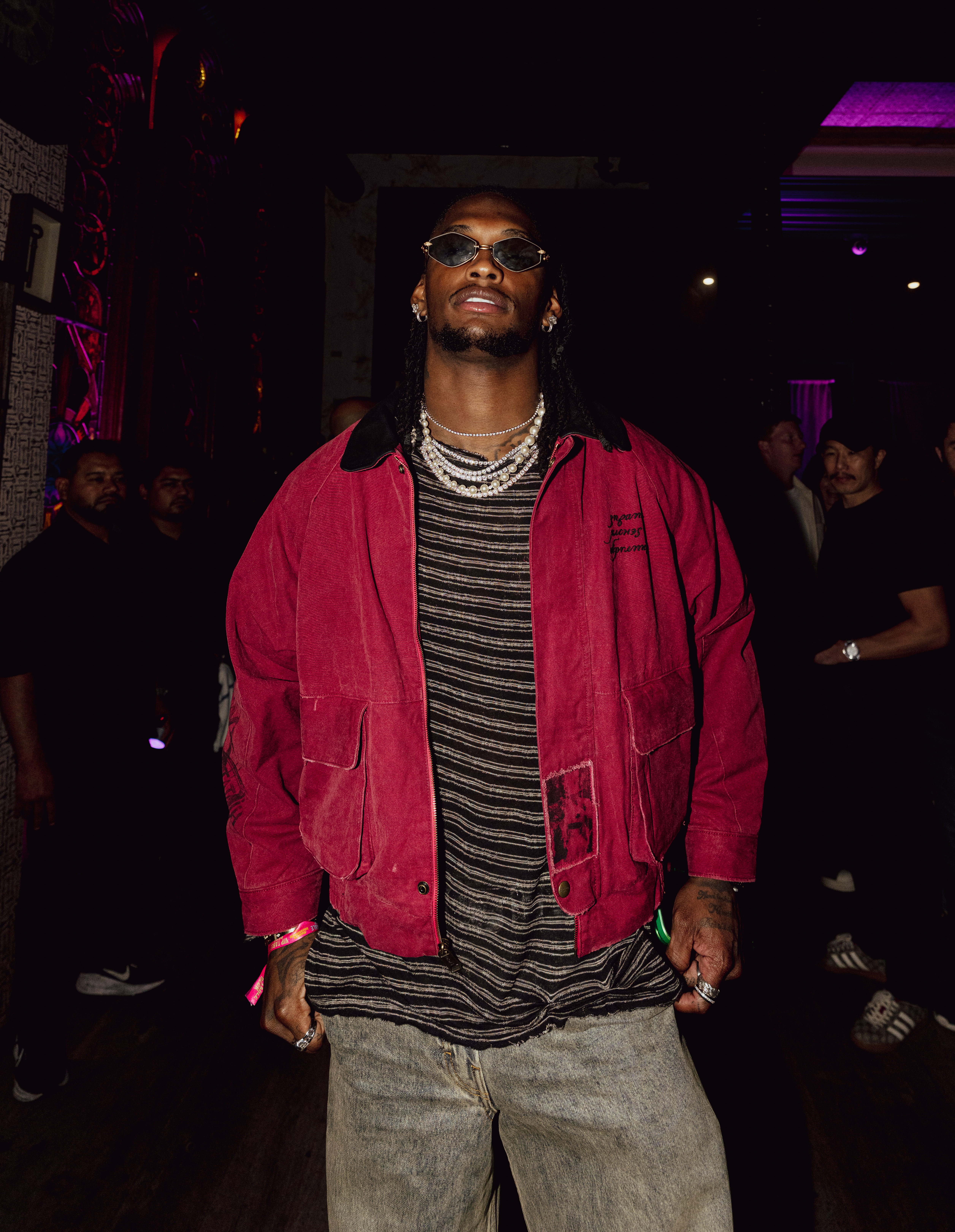 A man with dreadlocks wearing a red jacket, striped shirt, light-wash jeans, and sunglasses looks directly at the camera.