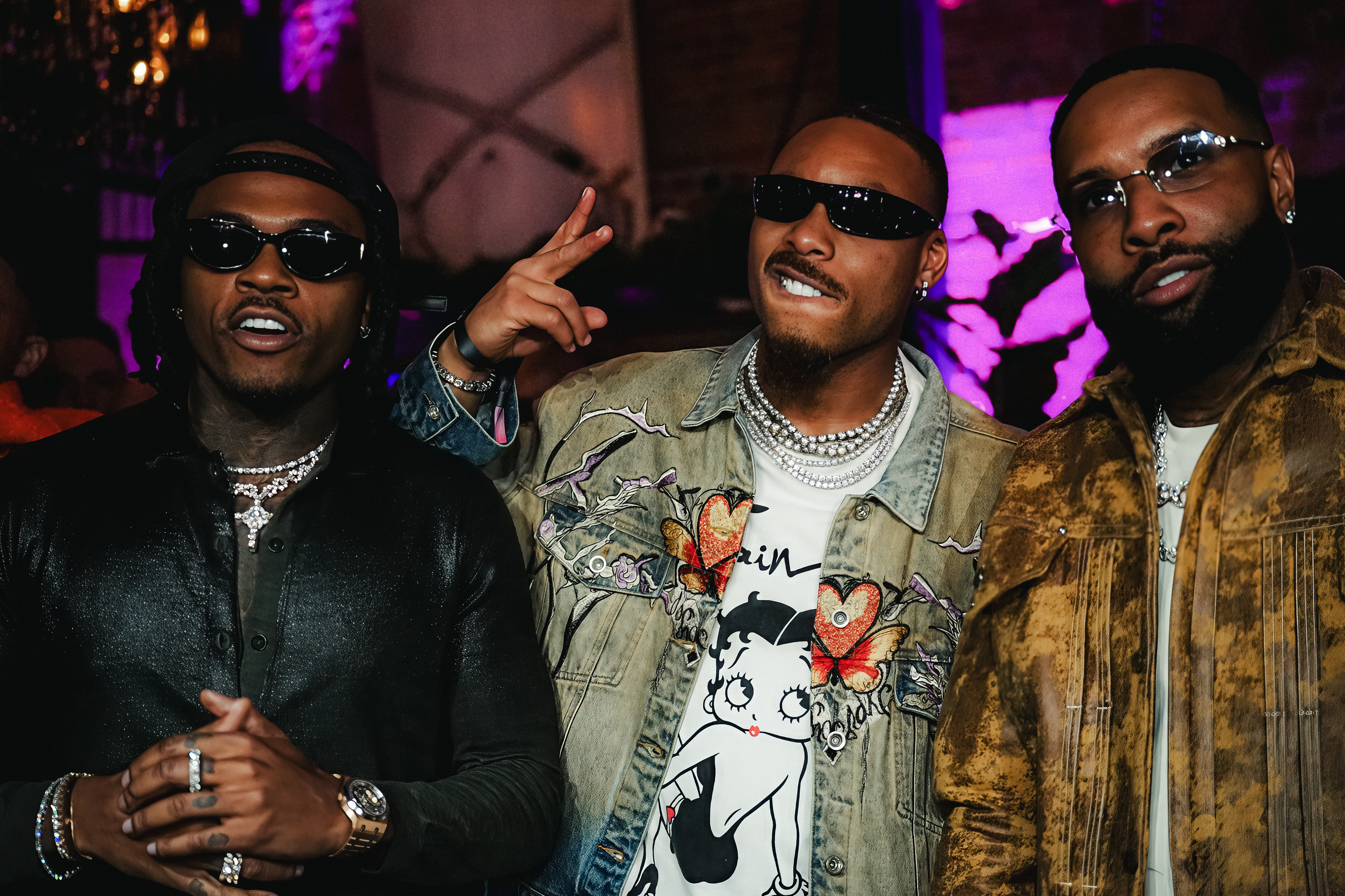 Three men in sunglasses, two of them smiling, standing in a dim room with purple lighting.