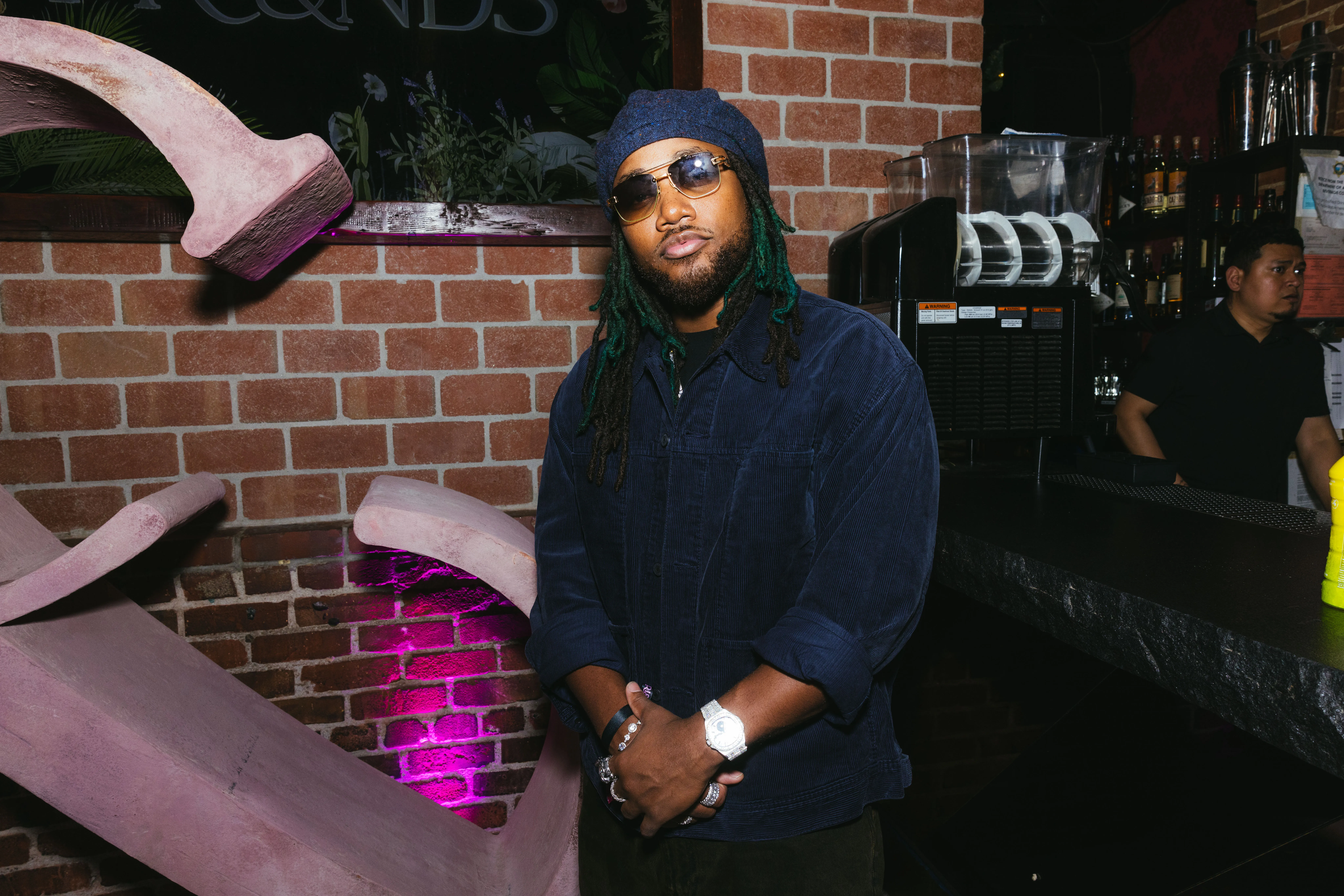 Leon Thomas in a dark blue corduroy jacket, black beanie, and sunglasses, posing indoors.