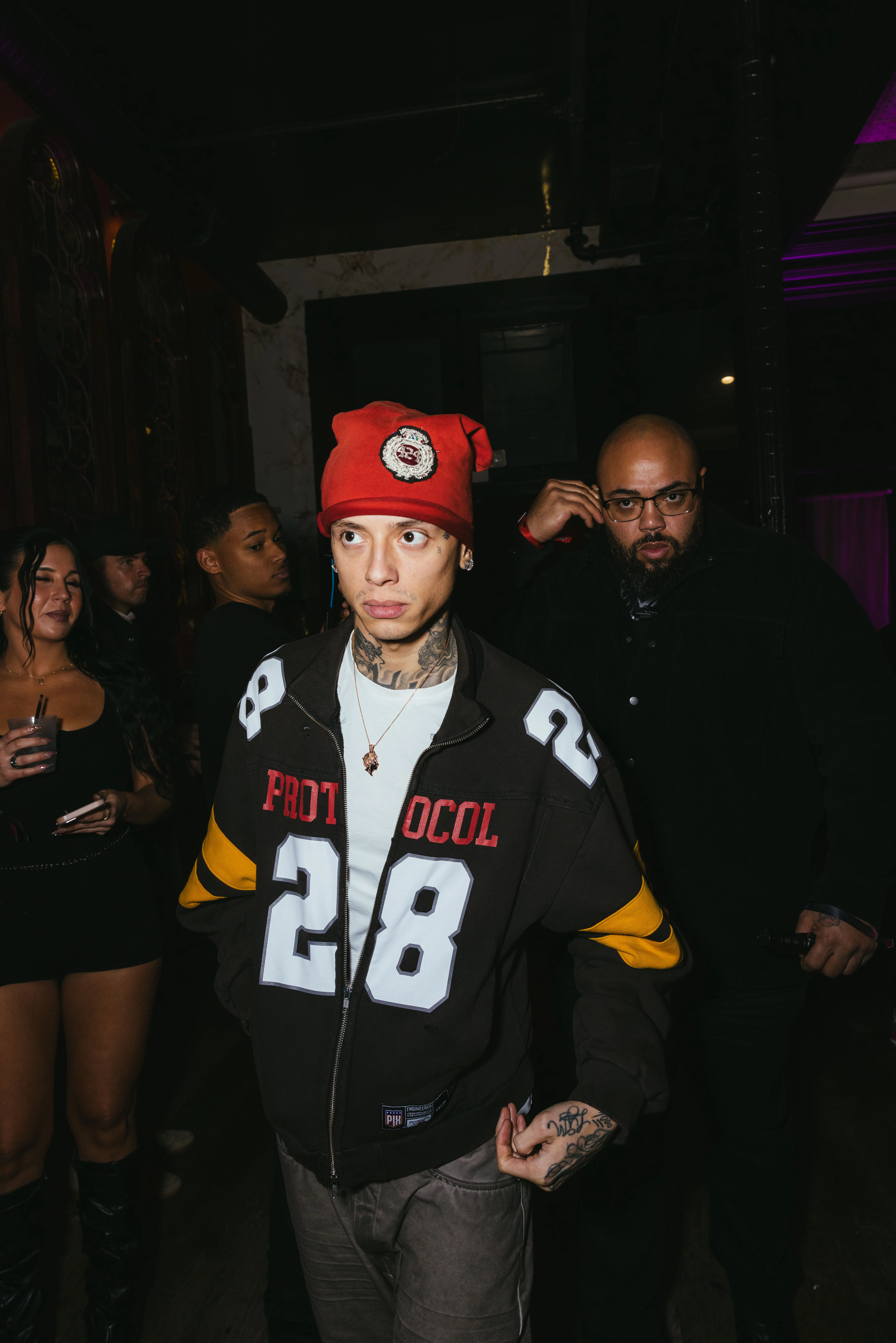 Rapper Central Cee in a red beanie and black and yellow jacket surrounded by people in a dimly lit venue.