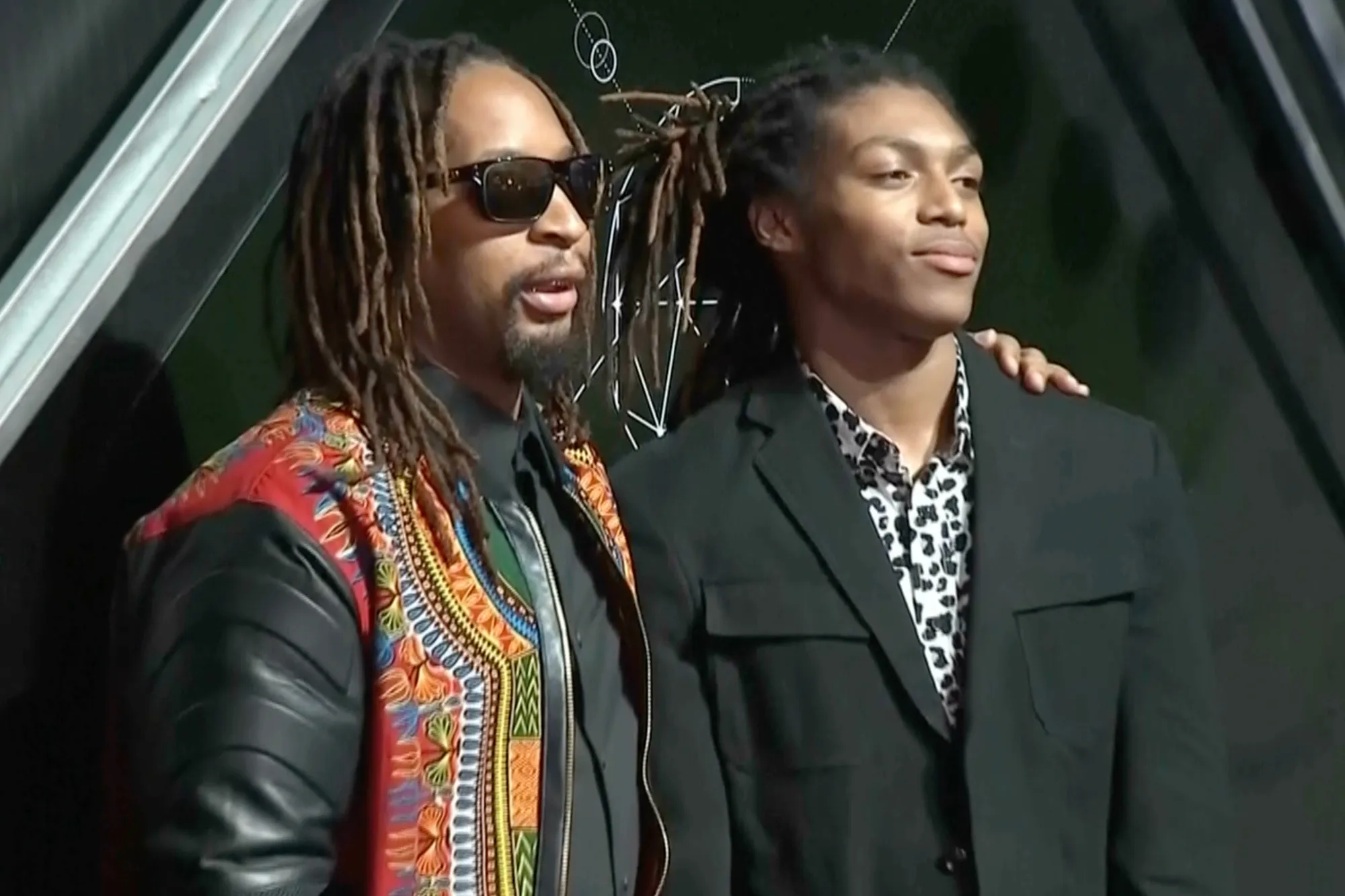 Man with dreadlocks and sunglasses in a patterned jacket standing next to a younger man in a dark suit.