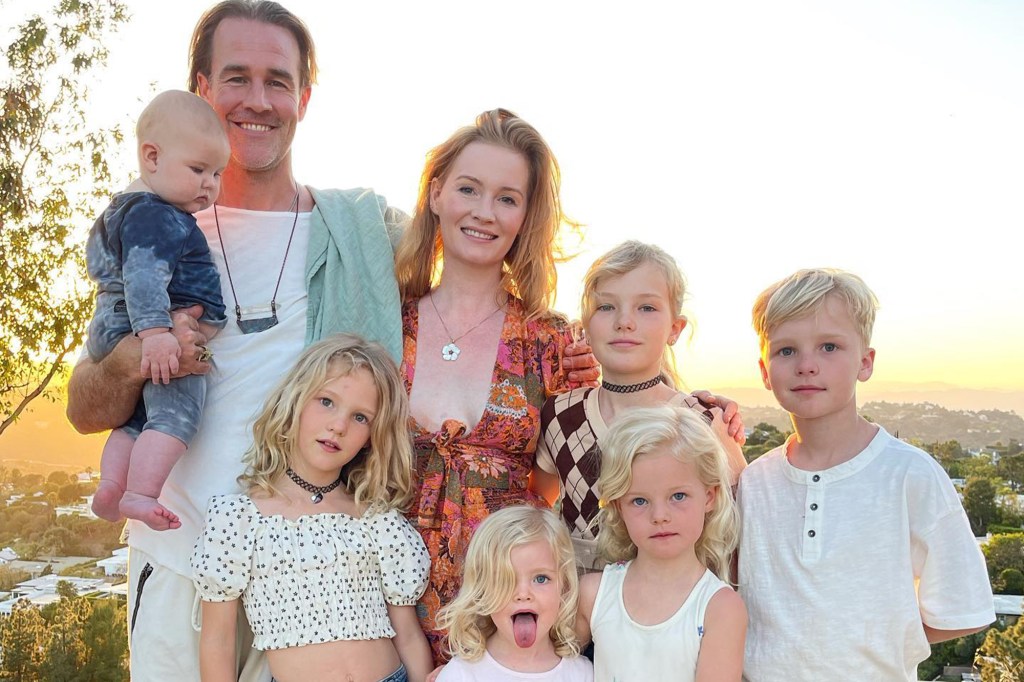 A family of eight, including six blonde-haired children and two adults, stands outdoors in golden hour light, smiling at the camera.