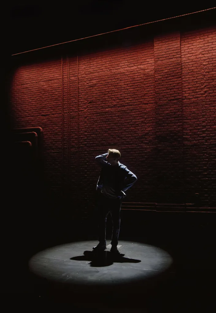 A man stands in a spotlight on a stage, illuminated from behind by a reddish light on a brick wall.