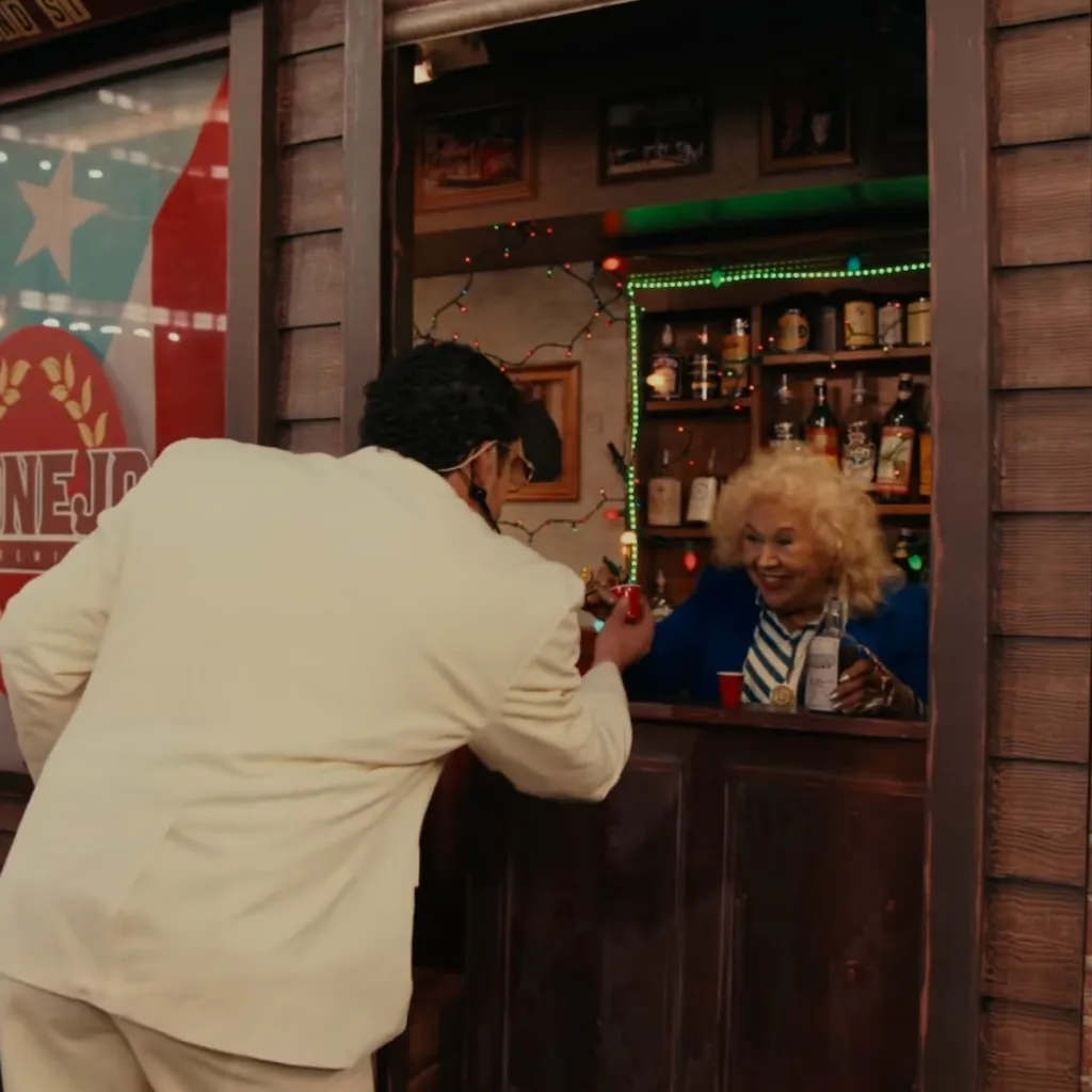 A man in a white suit leans into a bar window to get a shot from a smiling woman.