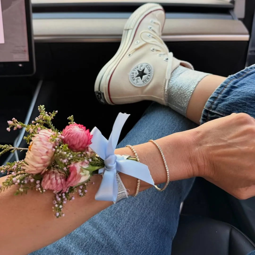 A person's arm wearing a corsage and diamond bracelet, with a Converse-clad foot resting in the background.