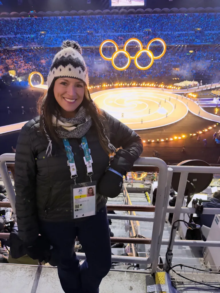 A woman in winter gear smiles in a stadium with the Olympic rings illuminated behind her.