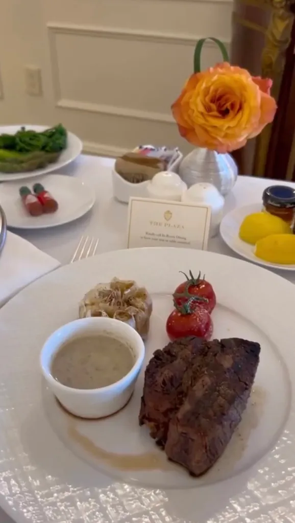A close-up of a steak dinner with roasted garlic, tomatoes, and a small bowl of sauce, served on a white plate with a card for 