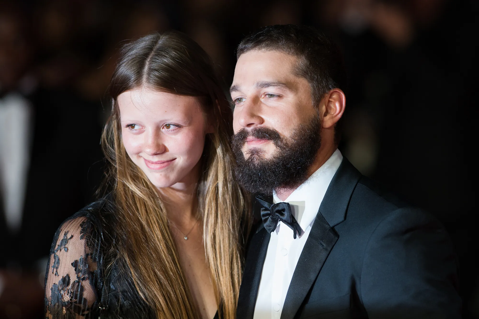 Shia LaBeouf, with a beard and wearing a bow tie, next to Mia Goth, both looking right.
