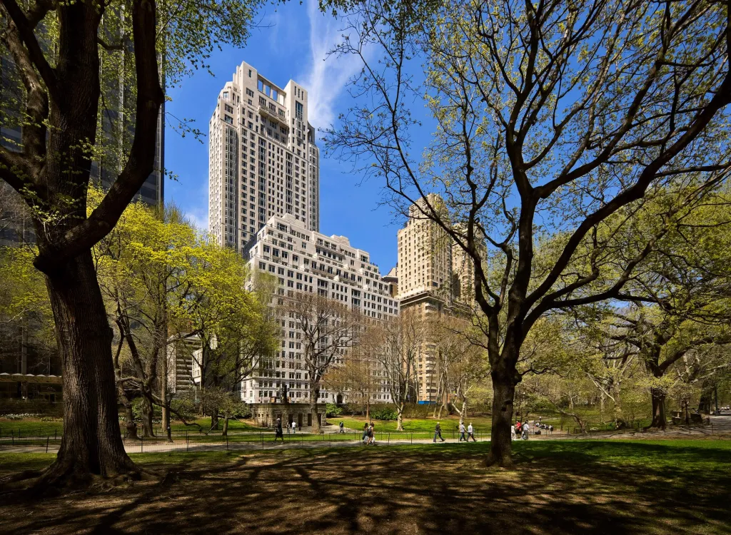 View from Central Park West, with a beige high-rise on the left and other buildings behind it, and lush green trees in the foreground.
