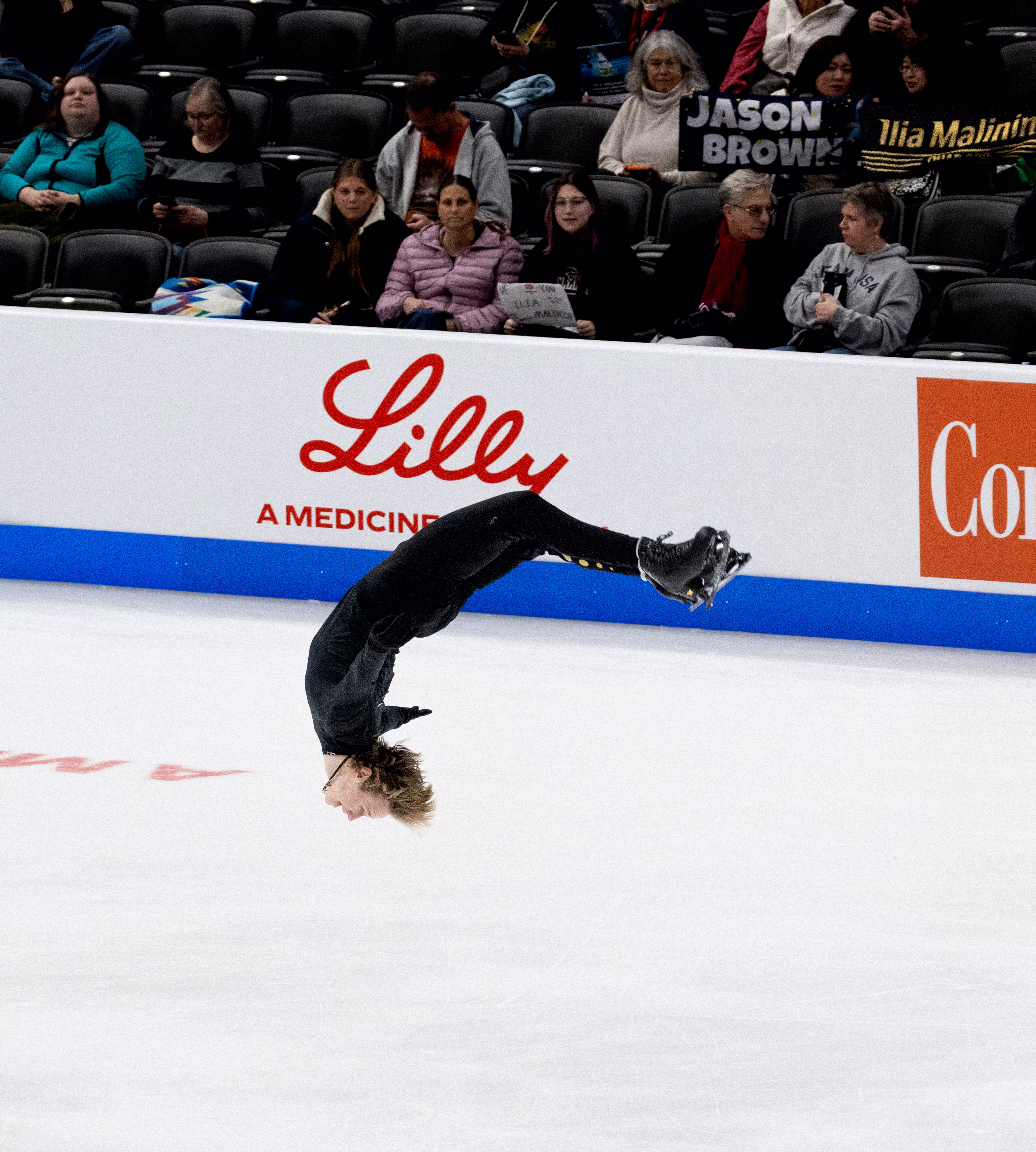 photo of ice rink with skater dressed in black in mid-flip upside down with legs and arms extended