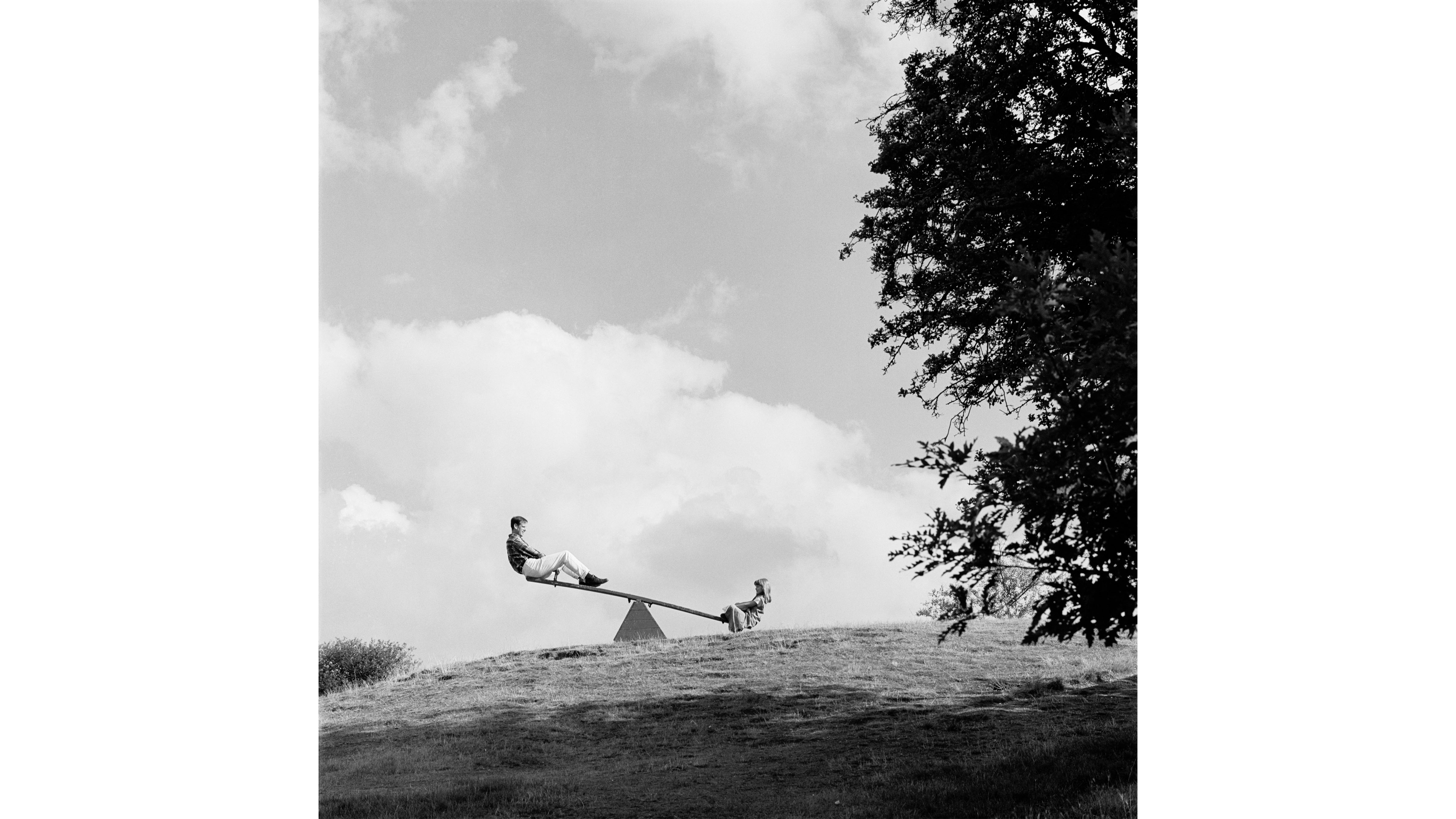 a black and white photograph of a father and daughter on a see saw next to a large tree