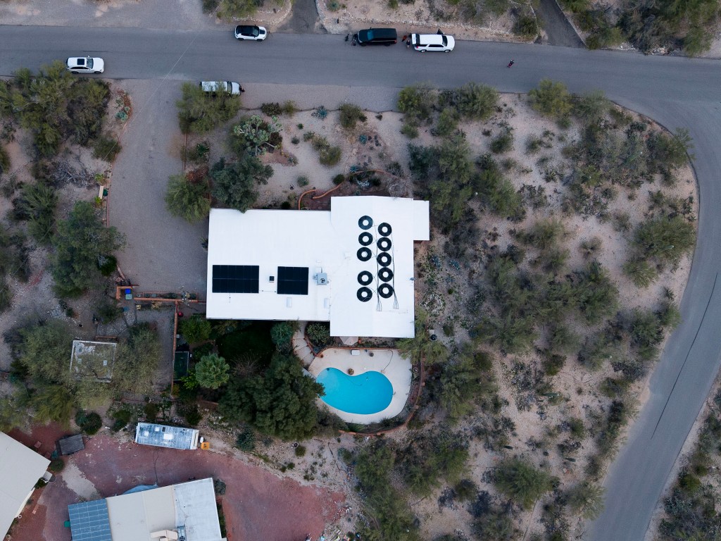 Aerial view of the home of Annie Guthrie and Tommaso Cioni in Tucson, AZ, showing a white roof with solar panels and a swimming pool, surrounded by desert landscaping and roads.
