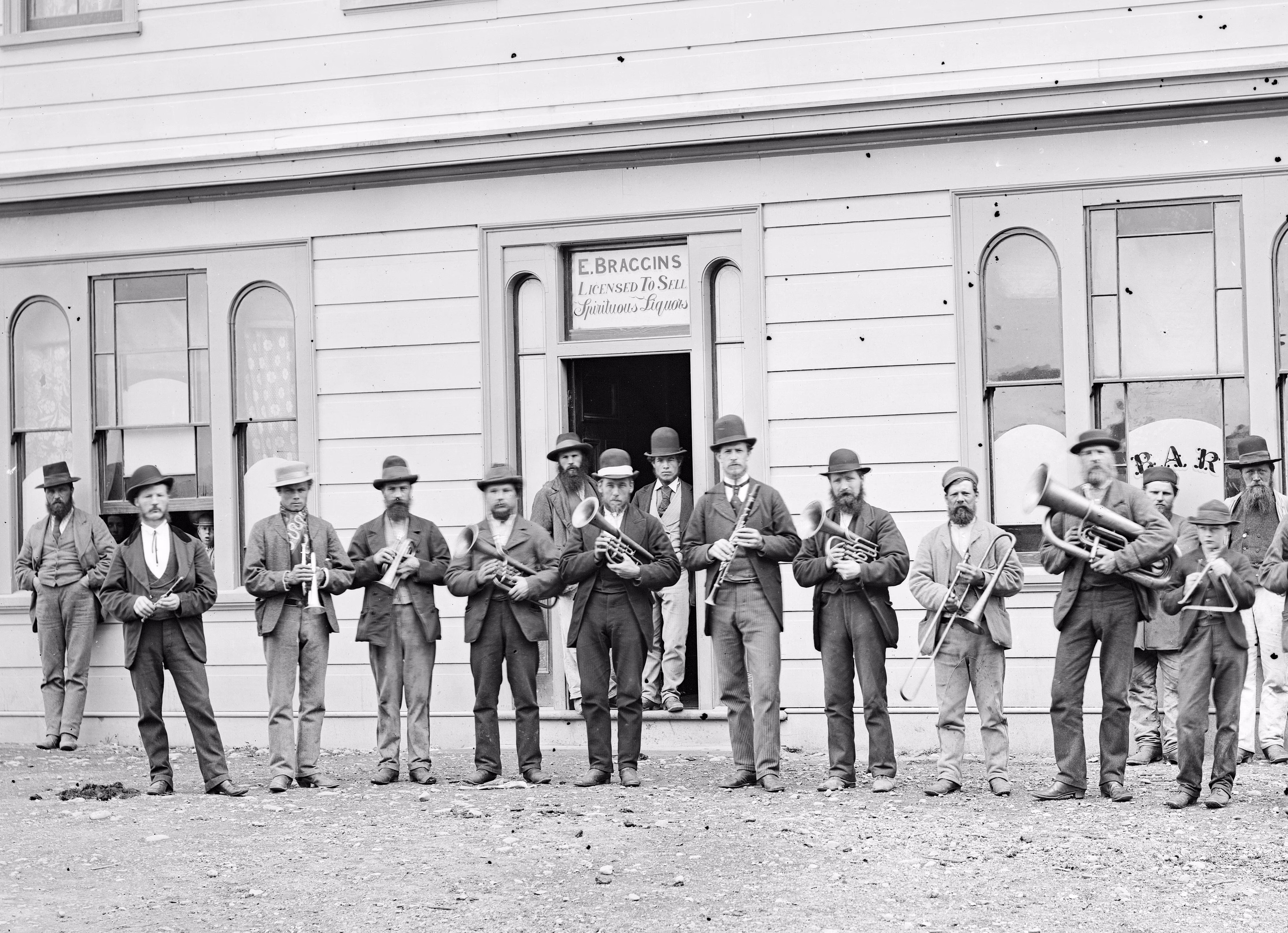 Members of a band pose with their instruments in front of a hotel, circa 1876. A sign above the door reads: “E. Braggins Licensed to sell spirituous liquors.”