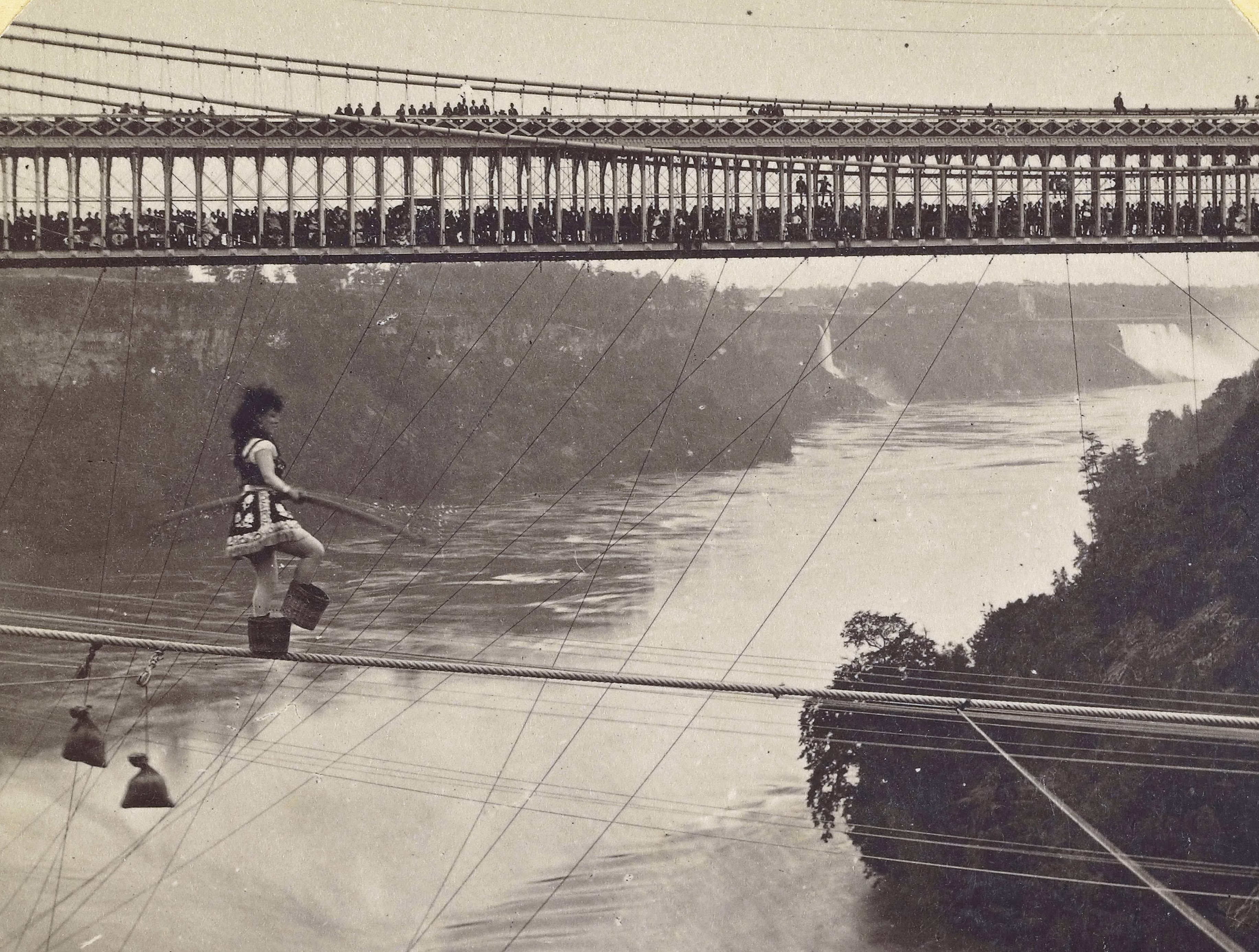 A large crowd of people look on from a bridge as a tightrope walker (wearing small baskets on their feet) walks on a rope above Niagara Falls.