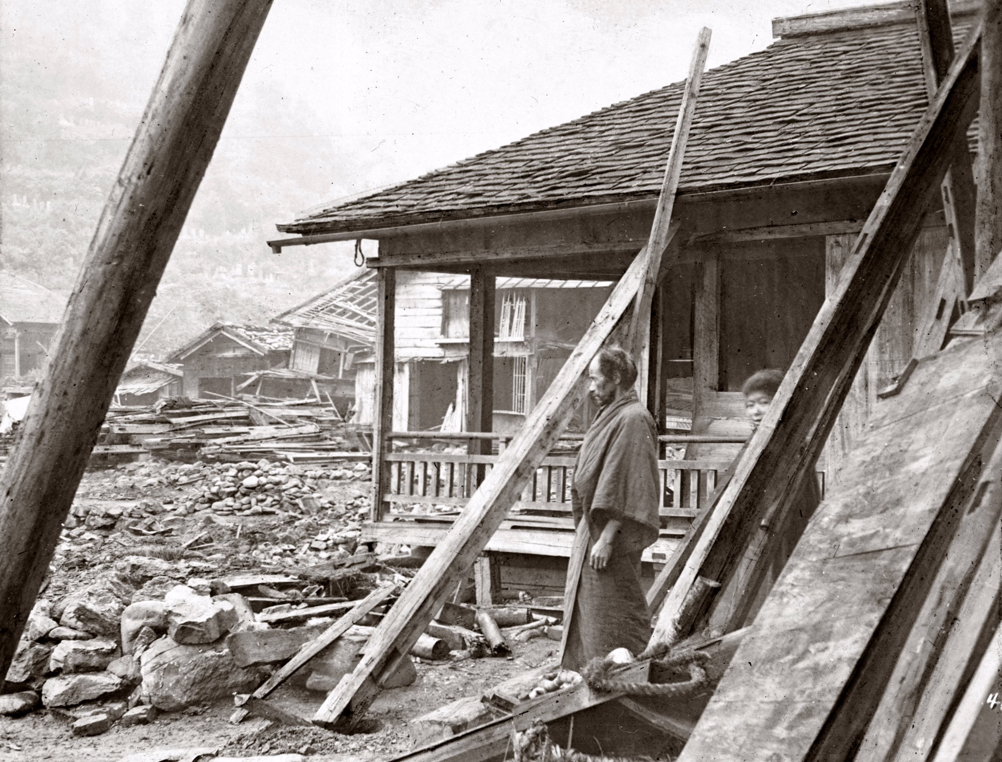 Two people stand outside a damaged house, looking at debris from other earthquake-damaged houses.