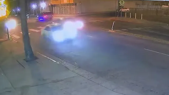 Security camera footage of a car with bright headlights driving down a street at night.