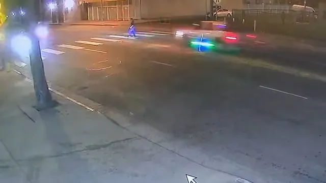 Night vision security footage of a pedestrian in a crosswalk as a vehicle with flashing blue and green lights approaches quickly.