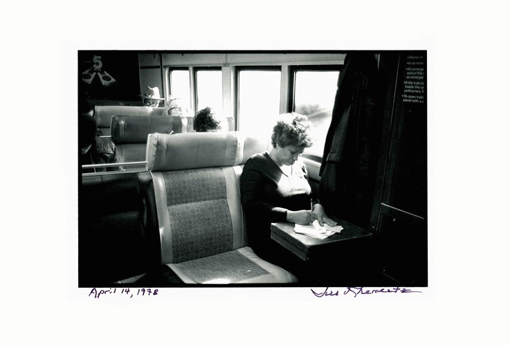 black-and-white photo of woman sitting in window seat on train, head bowed and writing on a briefcase on her lap, signed at bottom by photographer and hand-dated April 14, 1978