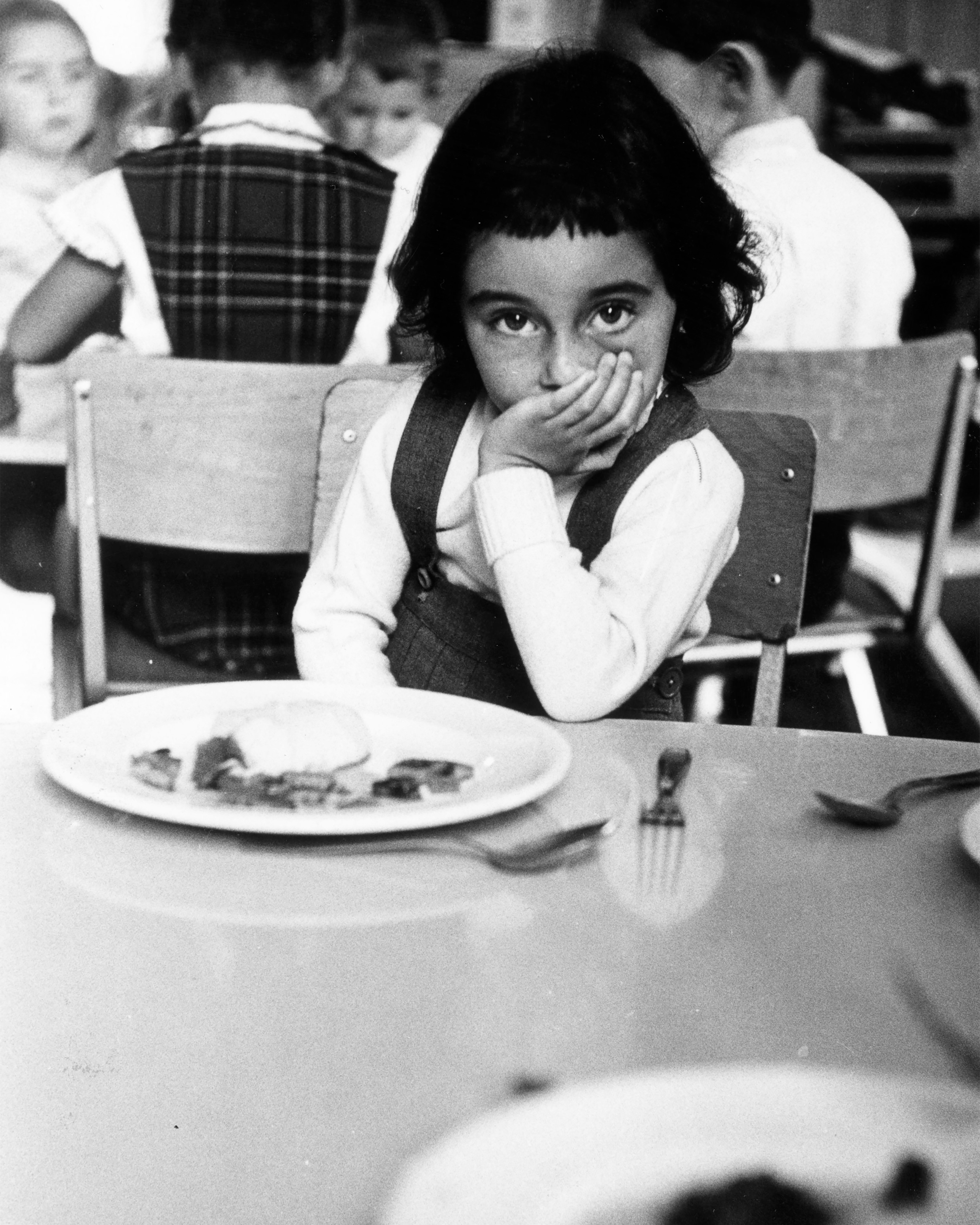 a child sitting with he hand on her mouth in front of a plate of food