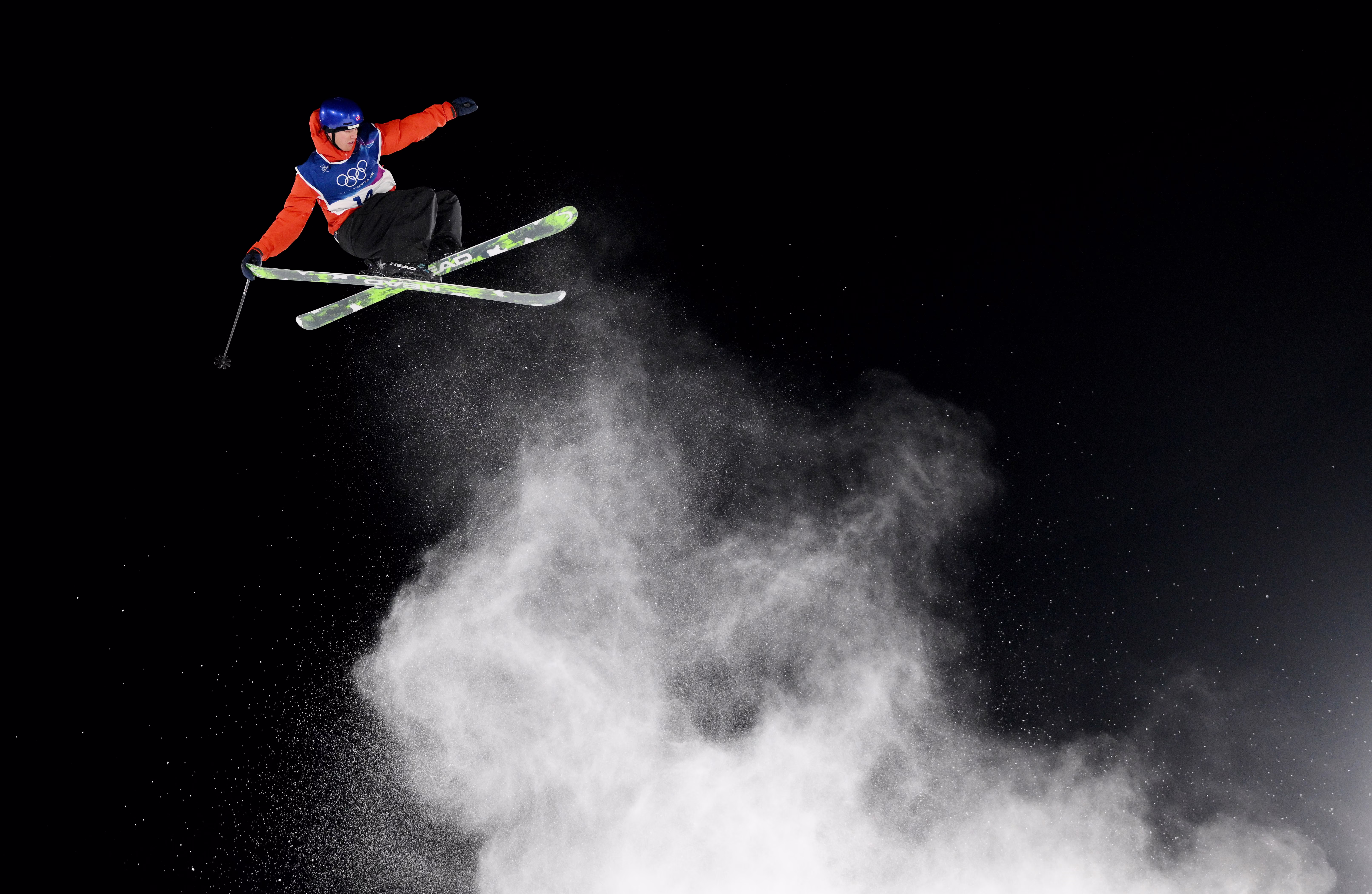 A skier launches from a jump, leaving a plume of flying snow, seen against a dark sky.