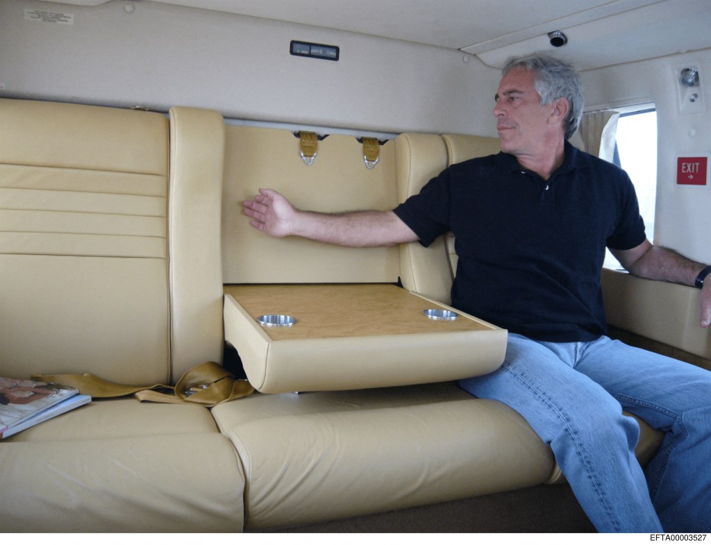 Jeffrey Epstein sitting on a cream-colored seat with a pull-down table and two cup holders, looking to his right.
