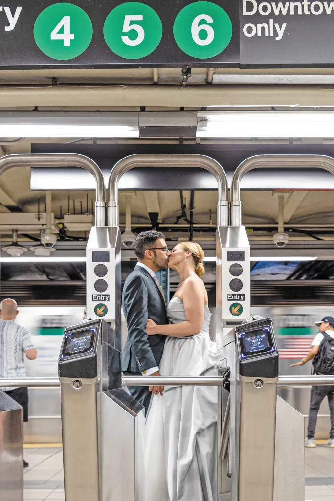 A couple in formal wear kissing between subway turnstiles, with a train blurred in the background.