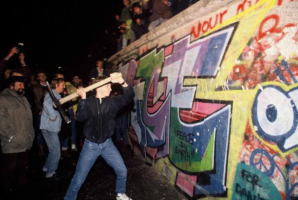A man attacks the graffiti-covered Berlin Wall with a pickaxe as a crowd watches.