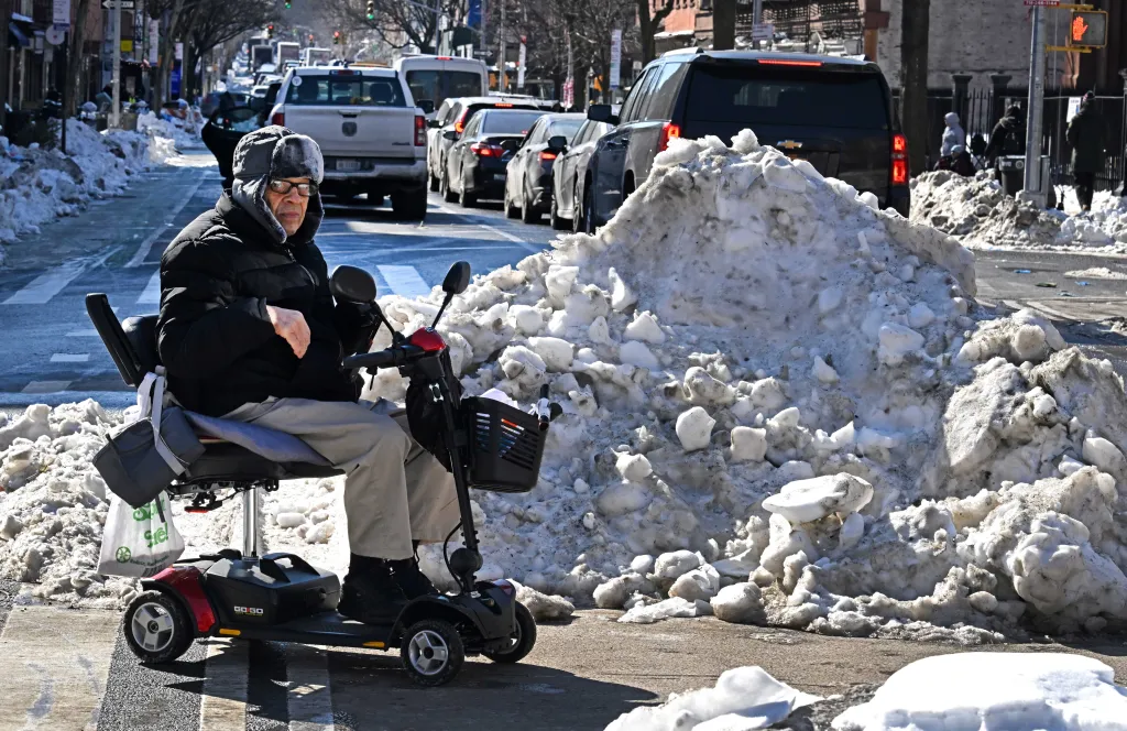 A man drives a motorized wheelchair past a large pile of snow on Court Street in Carroll Gardens. 