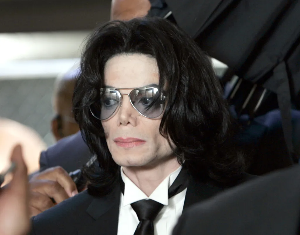 Michael Jackson, wearing aviator sunglasses, a white shirt, and a black suit and tie, looks to the right.