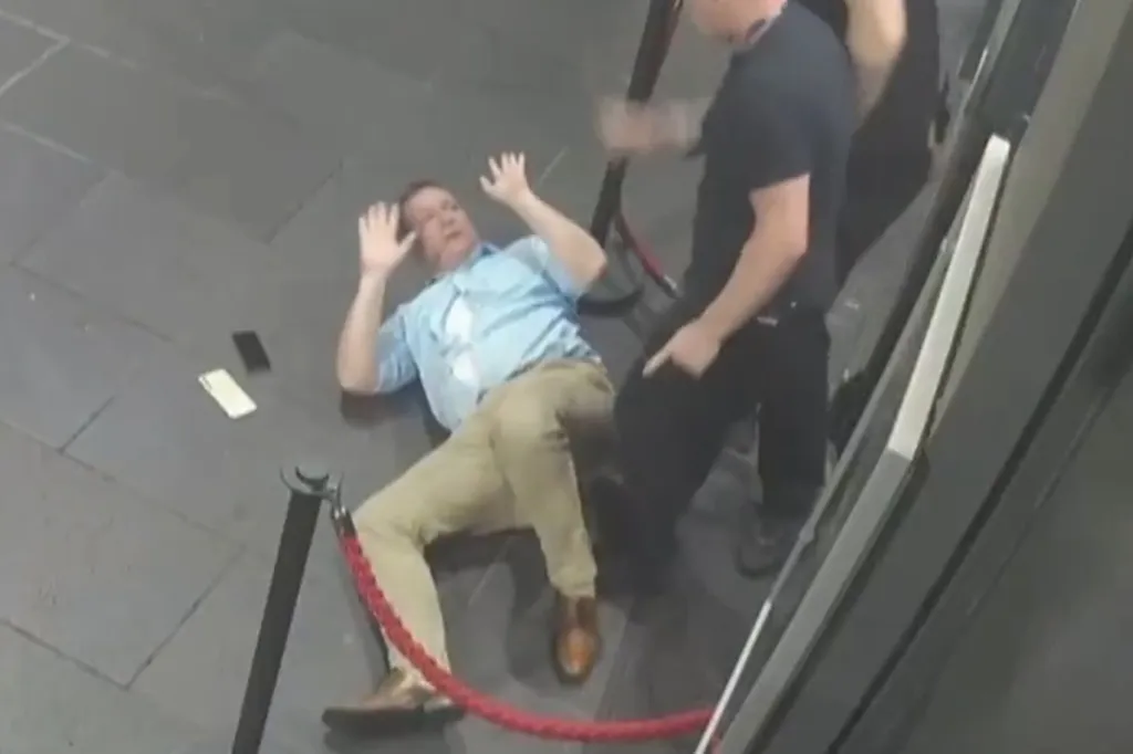 A man in a light blue shirt and khaki pants lies on his back on the floor with his hands raised, while another man in a black shirt stands over him.