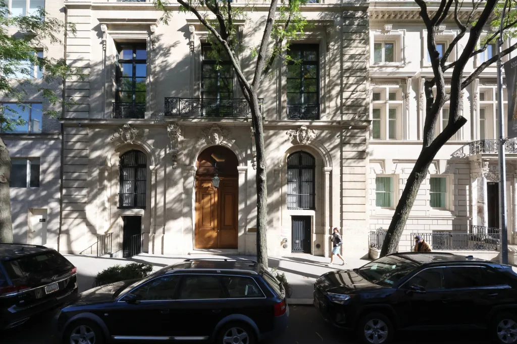 Exterior view of Jeffrey Epstein's former residence on East 71st Street, a large mansion with a prominent wooden door, flanked by two parked cars and shaded by trees.