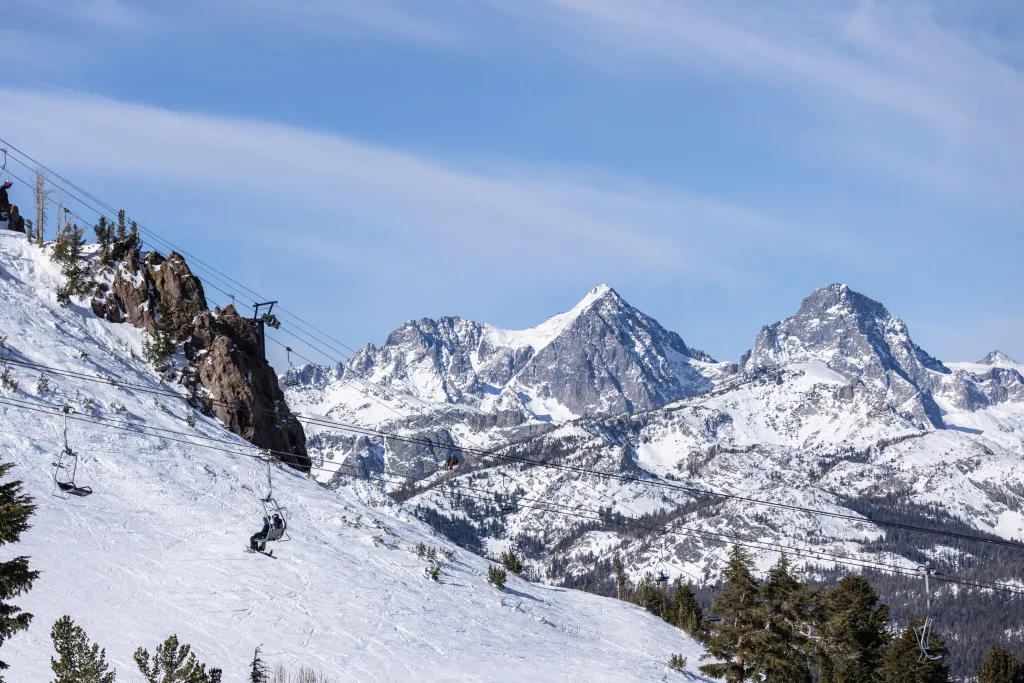 Skiers riding a chairlift up a snow-covered mountain with other peaks in the background.