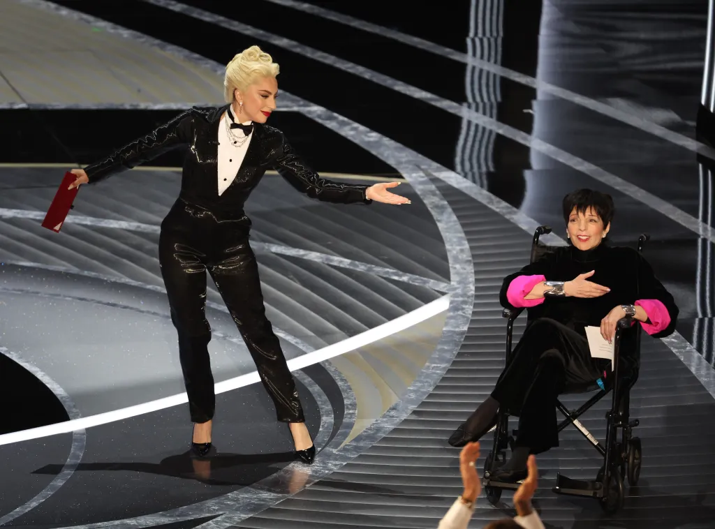 Lady Gaga in a shiny black tuxedo presenting an award to Liza Minnelli, who is seated in a wheelchair and wearing a black outfit with pink sleeves.