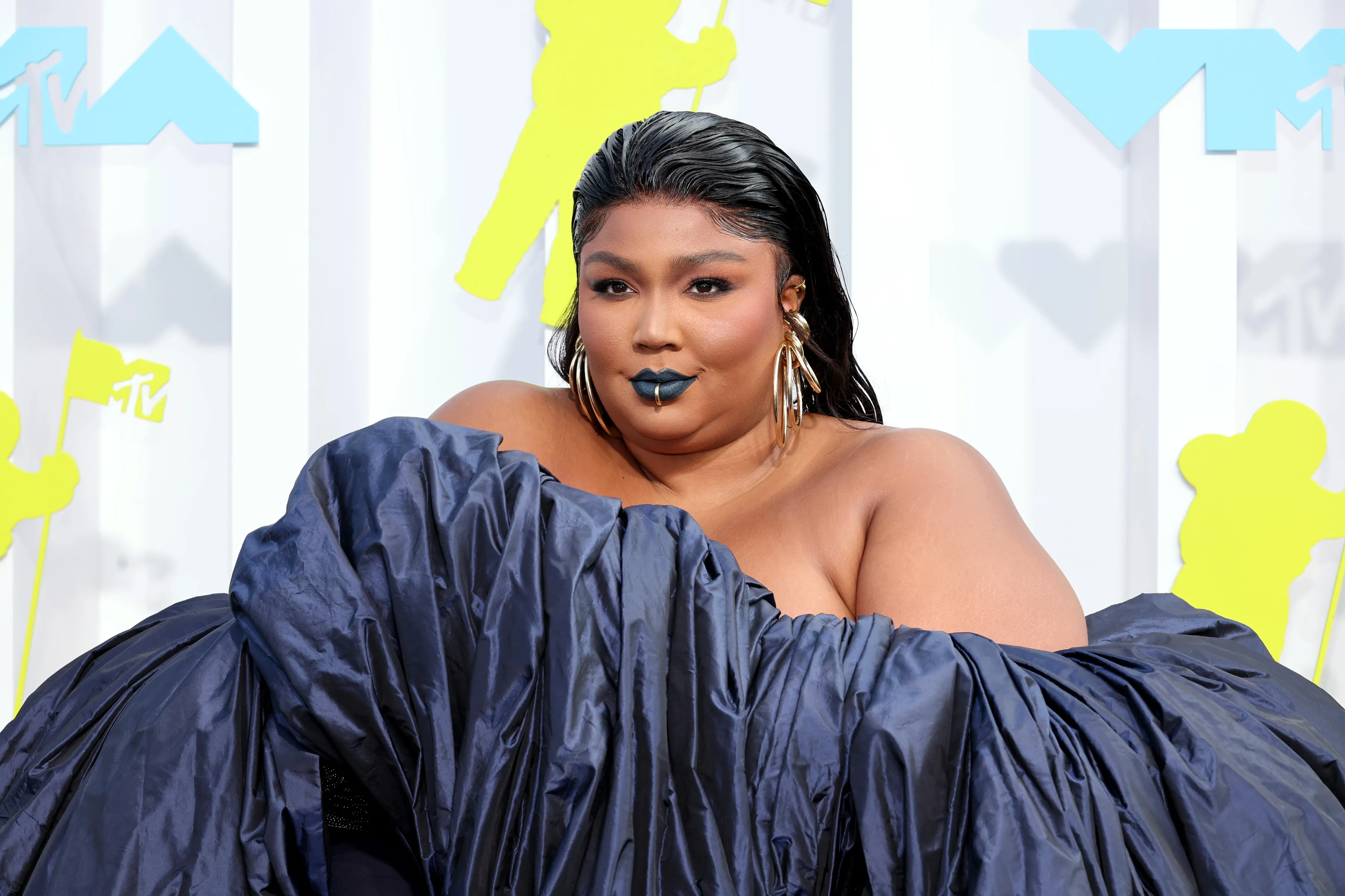 Lizzo in a dark blue off-the-shoulder gown, with slicked-back hair, dark blue lipstick, a gold lip ring, and gold earrings.