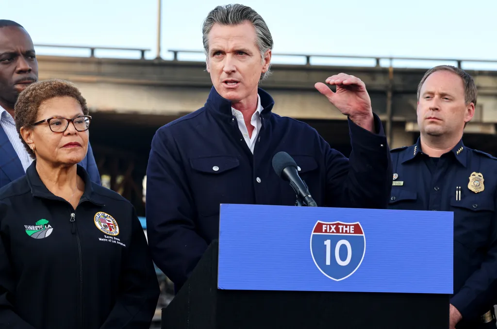 California Governor Gavin Newsom speaking at a podium with Los Angeles Mayor Karen Bass and a fire marshal, with an Interstate 10 sign and a damaged freeway in the background.