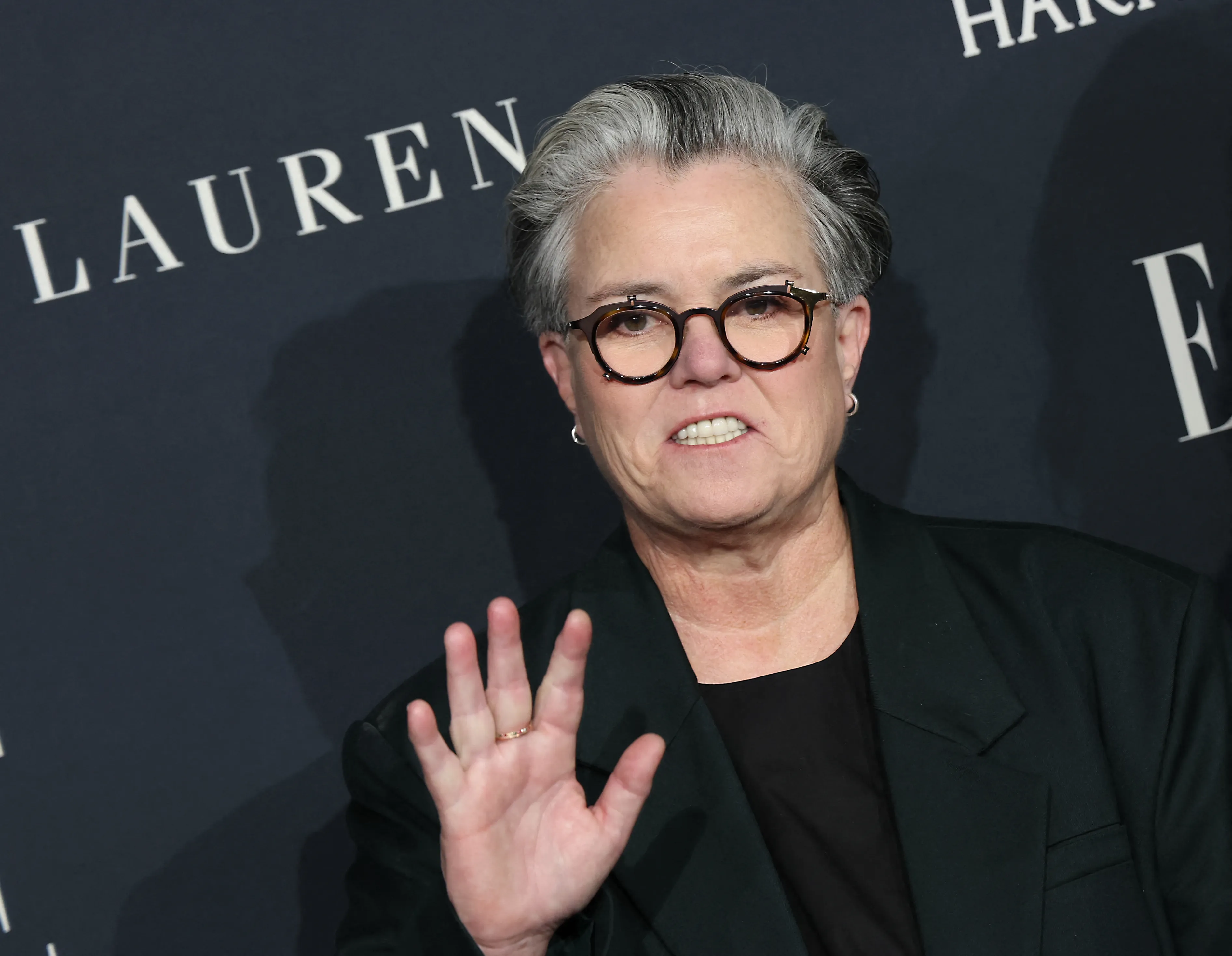 Rosie O'Donnell in a dark suit and glasses, waving her hand.