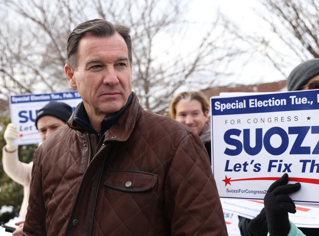 Tom Suozzi wearing a brown quilted jacket, with a campaign sign for Suozzi For Congress in the background.