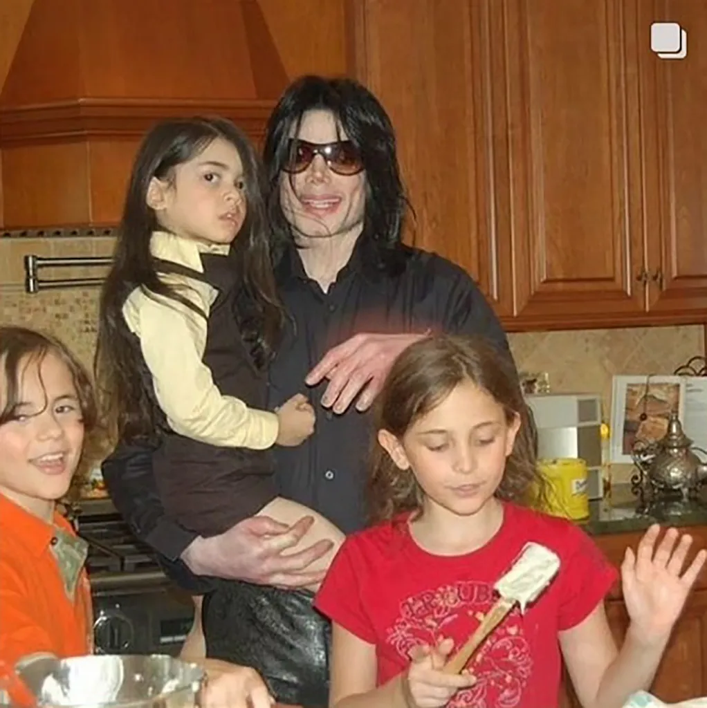 Michael Jackson, wearing sunglasses and a black shirt, holding young Paris Jackson, as other children gather around them in a kitchen.