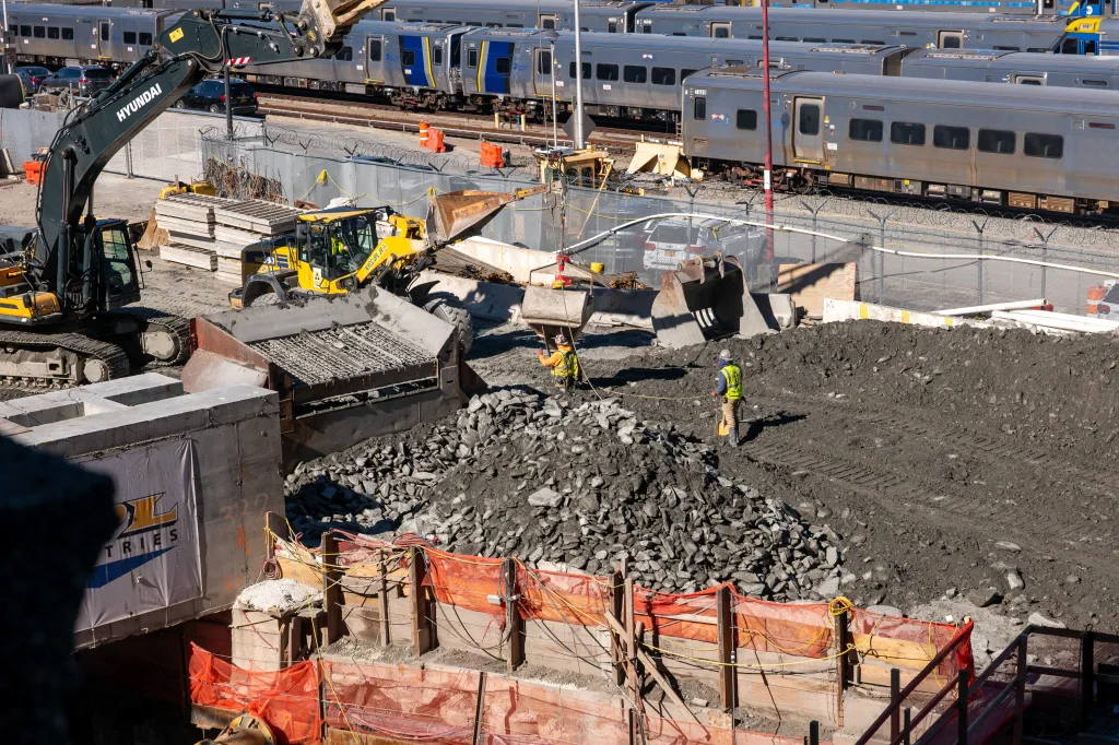 Construction on the Hudson Gateway Tunnel project, with a large excavator and workers in a dirt trench, and trains on tracks in the background.