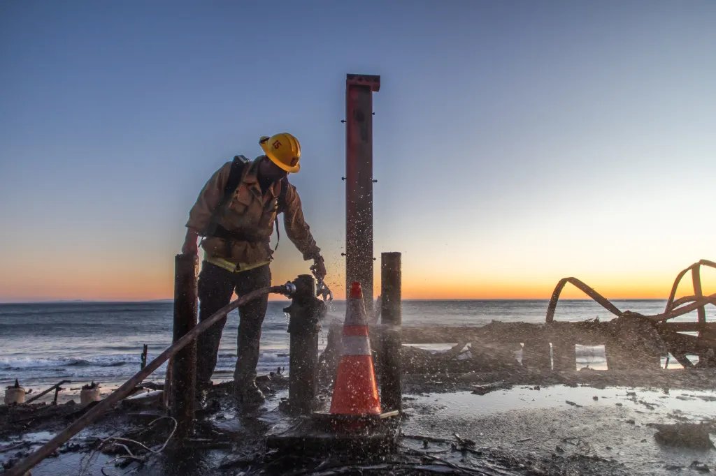 A firefighter working to switch off a fire hydrant on a wet, burnt landscape near the ocean at sunset.
