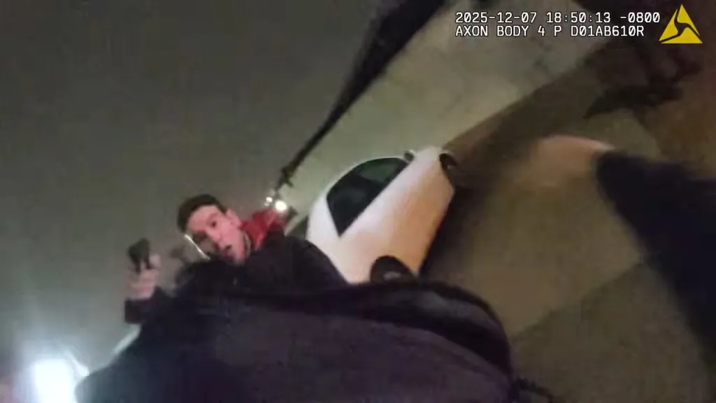 A still from a police bodycam video showing a man in a red shirt holding a gun while partially obscured by a car, looking towards the camera.