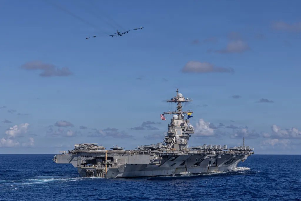 U.S. aircraft carrier USS Gerald R. Ford (CVN 78) at sea with three F/A-18E Super Hornets and a B-52H Stratofortress flying overhead.